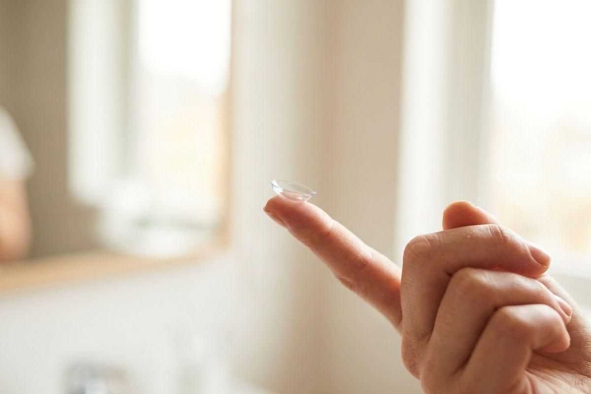 A contact lens balanced on the tip of a person's index finger, held up in a bright indoor setting.